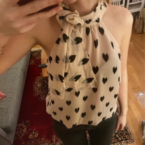 Beige ärmlös blus med hjärtmönster - Säljer en söt beige blus med svarta hjärtan och en knytning vid halsen. Den är ärmlös och har en luftig passform, perfekt för varmare dagar. 