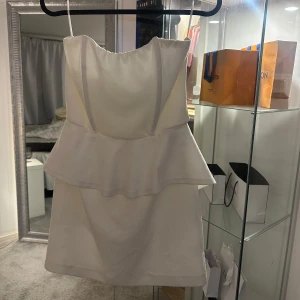 Vit axelbandslös klänning - Säljer en elegant vit axelbandslös klänning. Klänningen har en volangdetalj i midjan och en dragkedja bak. Perfekt för speciella tillfällen. Storlek S