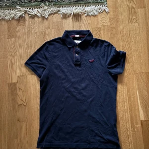 Mörkblå pikétröja från Hollister - Säljer en snygg mörkblå pikétröja från Hollister med korta ärmar och klassisk krage. Tröjan har en liten broderad logga på bröstet och knappar framtill. Perfekt för en avslappnad stil. 💙