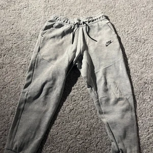 Grå mjukisbyxor från Nike - Säljer ett par grå mjukisbyxor från Nike med elastisk midja och snörning. Byxorna har en dragkedja på ena sidan och är perfekta för en avslappnad stil. Nike-loggan syns tydligt på framsidan. Strl 147-158