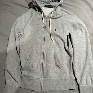 Grå hoodie från Polo Ralph Lauren - Säljer en stilren grå hoodie från Polo Ralph Lauren med dragkedja och justerbar huva. Tröjan har två praktiska fickor framtill och en liten broderad logga på bröstet. Knappt täcken på användning, väldigt sparsamt använd, inte noppring eller något slitage vid muddar osv. 