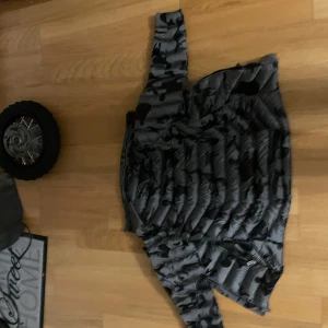 Grå camouflage dunjacka från Stone Island - Säljer en grå dunjacka från Stone Island med ett coolt camouflage-mönster. Jackan har en dragkedja framtill och är perfekt för kyligare dagar. Den är långärmad och har en modern look. Passar dig som vill ha något stilrent och funktionellt. Förlåt om bilderna är upp och ner. Har tyvärr inte kvittot och lådan men om du scannar qr koden så är den äkta 