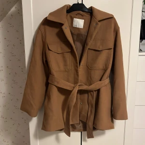 Brun jacka  - Snygg brun jacka från Samsa Collection med knytbälte i midjan och långa ärmar. Jackan har en klassisk krage och två stora fickor framtill. Perfekt för höst och vår.