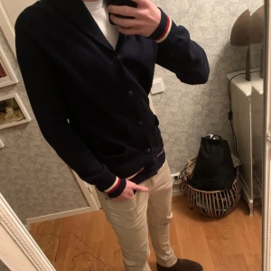 Mörkblå kofta från Polo Ralph Lauren - Säljer en snygg mörkblå kofta från Polo Ralph Lauren med knappar och ribbade detaljer. Den har vita och röda ränder vid ärmslut och nederkant. Perfekt för en stilren look.