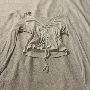 Vit ribbad topp med knytning - Säljer en stilren vit ribbad topp med lång ärm och knytning framtill. Toppen har en v-ringad design och är croppad för en modern look. Perfekt för att styla med jeans eller kjol.