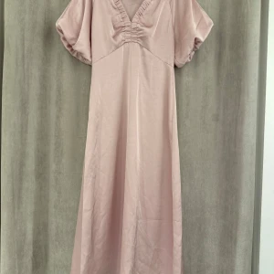 Neo noir klänning - Elegant ljusrosa långklänning med puffiga kortärmar och v-ringad design. Klänningen har en mjuk och flödande siluett som ger en romantisk känsla. Perfekt för speciella tillfällen.