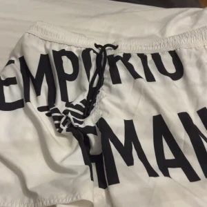 Vita badshorts från Emporio Armani - Snygga vita badshorts från Emporio Armani med svart text och snörning i midjan. Perfekta för en dag på stranden eller vid poolen. Stilren design med en bekväm passform.