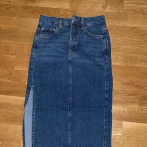 Blå jeanskjol med slits - Snygg blå jeanskjol med en stilren design och en slits på sidan. Perfekt för en avslappnad look. Kjolen har klassiska jeansfickor och bälteshällor.