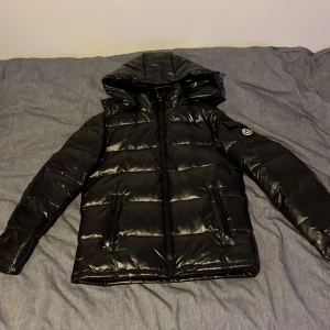 Svart pufferjacka med huva - Snygg svart pufferjacka från fashion med huva och dragkedja. Jackan har en glansig finish och långa ärmar. Perfekt för kyligare dagar när du vill hålla stilen. Helt oanvänd och priset kan diskuteras så slå en pling om du vill köpa 👍