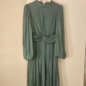 Grön långklänning med plisserade detaljer - Elegant grön långklänning med plisserade detaljer och långa ärmar. Klänningen har en hög hals och ett vackert knytband i midjan som ger en smickrande silhuett. Perfekt för speciella tillfällen.