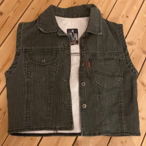 Grön jeansväst från ACM - Säljer en cool grön jeansväst från ACM med knappar framtill och två bröstfickor. Västen har en klassisk krage och är perfekt för en avslappnad stil. Passar bra över en t-shirt eller skjorta. Bra skick!