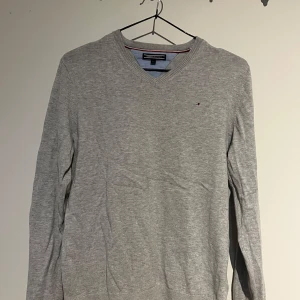 Grå tröja från Tommy Hilfiger - Säljer en stilren grå tröja från Tommy Hilfiger med v-ringning och diskret logga på bröstet. Tröjan har långa ärmar och är tillverkad i ett mjukt material som passar perfekt för kyligare dagar.