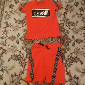 Orange t-shirt och shorts från Cavalli - Snyggt set med orange t-shirt och matchande shorts från Cavalli. T-shirten har korta ärmar och ett stort Cavalli-tryck på bröstet. Shortsen har svarta detaljer längs sidorna och en bekväm resår i midjan.  Den har lite fläckar men märks bara när man tittar nära.
