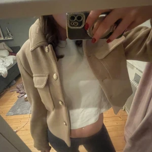 Beige kappa från Zara - Snygg beige jacka från Zara med knappar och två stora fickor framtill. Jackan är croppad och har en klassisk krage. Helt oanvänd med prislapp kvar