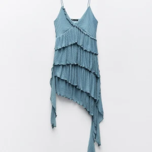 Blå asymmetrisk klänning med volanger - Snygg blå klänning med smala axelband och asymmetrisk design. Klänningen har flera lager av volanger som ger en unik look. Perfekt för en somrig dag eller en speciell kväll. Aldrig använd. Prislapp kvar