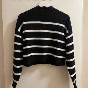 Svart och vit randig stickad tröja - Säljer en snygg svart och vit randig stickad tröja med långa ärmar. Tröjan har en hög krage och är perfekt för kyligare dagar. Den har en croppad passform som ger en modern look.