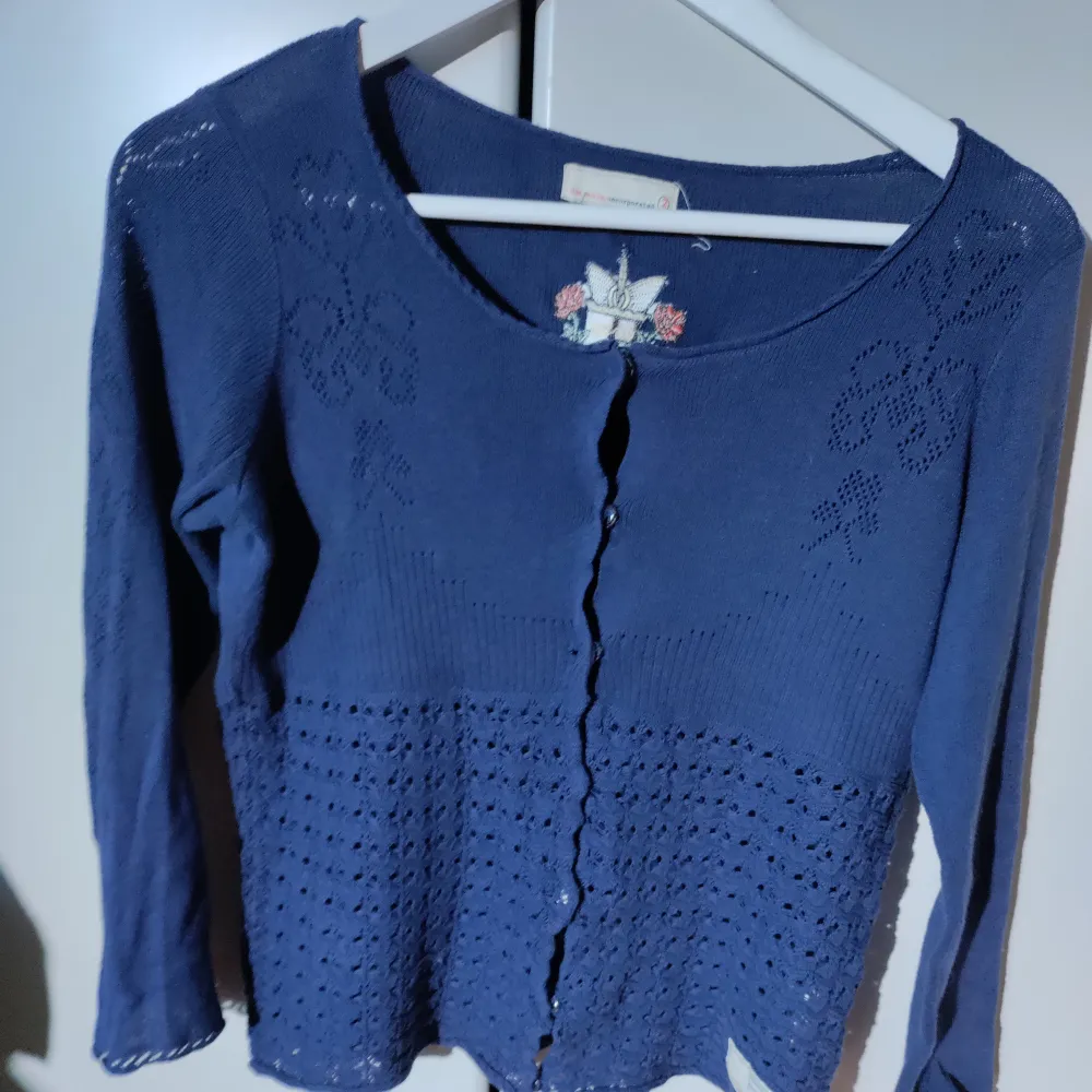 Säljer en mörkblå kofta med vackert broderi på ryggen. Koftan har långa ärmar och knappar framtill. Perfekt för en stilren look.. Neuletakit & Villapaidat.