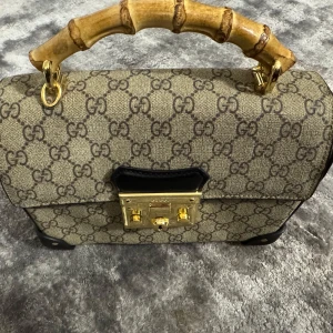 Mönstrad handväska med bambuhandtag - Snygg handväska med klassiskt mönster och unikt bambuhandtag. Väskan har en elegant låsdetalj i guld och är perfekt för att ge en touch av lyx till din outfit.