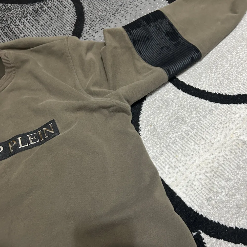 Snygg olivgrön tröja från Philipp Plein med logotyp framtill. Tröjan har långa ärmar med svarta detaljer vid armbågarna. Perfekt för en stilren look.. Neuletakit & Villapaidat.