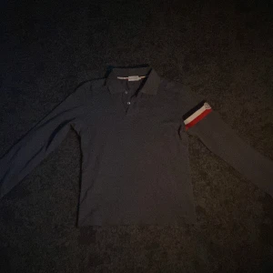 Grå pikétröja från Moncler - Snygg grå pikétröja från Moncler med långa ärmar och klassisk krage. Tröjan har en röd och vit randdetalj på ärmen med Moncler-logotypen. Perfekt för en stilren look.