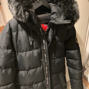 Svart dunjacka med pälsdetaljer från Zavetti Canada - Snygg svart dunjacka från Zavetti Canada med en fluffig pälsdetalj på huvan. Jackan har en dragkedja framtill och ett stilrent märke på ärmen. Perfekt för kalla vinterdagar.