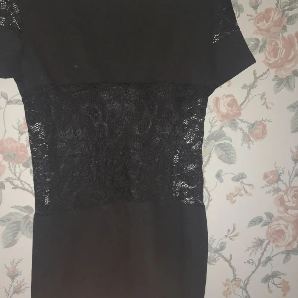 Elegant svart klänning med spetsdetaljer på axlar och sidor. Klänningen har korta ärmar och en smickrande passform. Perfekt för en stilren look.. Mekot.