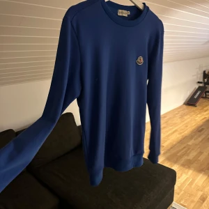 Blå tröja från Moncler - Säljer en stilren blå tröja från Moncler med rund hals och ett litet broderat märke på bröstet. Tröjan har långa ärmar och är perfekt för en avslappnad look. Passar bra till både jeans och chinos.