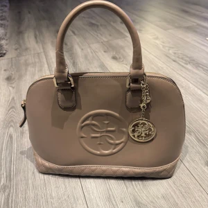 Beige handväska från Guess - Snygg beige handväska från Guess med quiltad botten och ett elegant metallmärke. Väskan har dubbla handtag och en dragkedja upptill. Perfekt för att ge en stilren touch till din outfit.