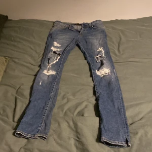 Blå slitna jeans från Jack & Jones - Säljer ett par blå jeans från Jack & Jones med slitna detaljer och hål på benen. De har en klassisk femficksdesign och knappgylf. Perfekta för en avslappnad stil.