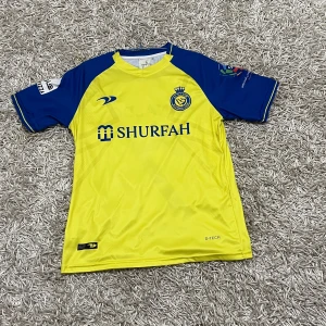 Fotbollströja Al-Nassr - Säljer en gul och blå fotbollströja från D-Tech med korta ärmar. Tröjan har ett tryck med texten 'SHURFAH' på framsidan och 'RONALDO 7' på baksidan. Perfekt för fotbollsfans!
