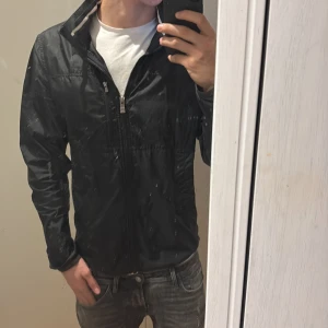 Svart vindjacka från J.Lindeberg - Snygg svart vindjacka från J.Lindeberg med dragkedja och hög krage. Perfekt för blåsiga dagar och enkel att matcha med olika outfits. Jackan har en stilren design och är tillverkad i ett lätt material. Har en luva man kan väckla ut när det regnar.