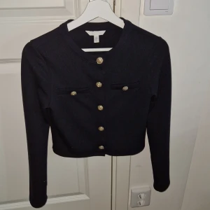 Svart tröja från H&M - Snygg svart tröja från H&M med guldknappar. Den har en croppad design och långa ärmar, perfekt för en stilren look. Tröjan  är i en klassisk stil med dekorativa knappar framtill.