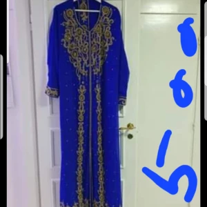 Blå långklänning med guldbroderi - Elegant blå långklänning med detaljerat guldbroderi längs framsidan och runt halsen. Klänningen har långa ärmar och en vacker, flytande design som ger en lyxig känsla.