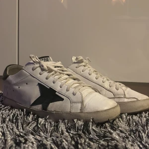 Vita  Golden Goose - Snygga vita sneakers från Golden Goose med en svart stjärndetalj på sidan. Skorna har snörning och en svart häl med märkeslogga. Perfekta för en avslappnad stil. St:42 passar 43. Cond: 6/10 har kvar att ge har heeldrag på båda skorna! Skriv om du vill ha fler bilder på sula etc