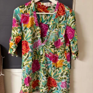 Blommig klänning från H&M - Säljer en färgglad blommig klänning från H&M med v-ringning och knappar framtill. Klänningen har trekvartsärmar och ett livfullt mönster i rosa, orange och grönt.