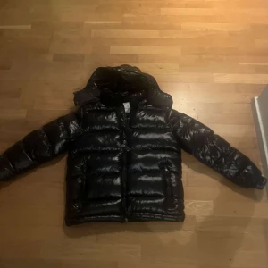 Svart dunjacka från Moncler - Säljer en snygg svart dunjacka från Moncler med huva och dragkedja. Jackan har en glansig finish och är perfekt för kyliga dagar. Den har långa ärmar och en klassisk pufferdesign.