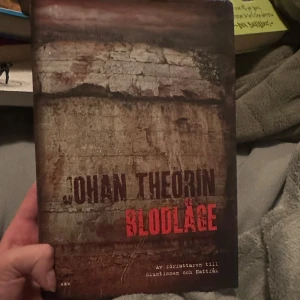 Blodläge - Boken 'Blodläge' av Johan Theorin har ett omslag med en dramatisk bild av en klippa i jordnära toner. Titeln är tryckt i rött och författarens namn i mörkare nyanser. Denna bok är en del av en serie och är känd för sin spännande handling.