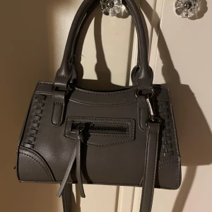 Grå handväska med dragkedja - Snygg grå handväska med dekorativa sömmar och en praktisk dragkedja framtill. Väskan har dubbla handtag och en avtagbar axelrem. Sparsamt använd så i fint skick! 