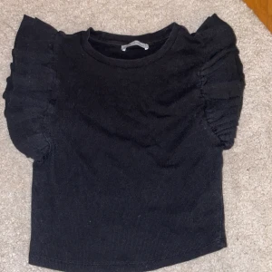 Svart topp med volangärmar från Zara - Snygg svart topp från Zara med volangdetaljer på ärmarna. Perfekt för att ge en extra touch till din outfit. Toppen har en rund halsringning och är tillverkad i ett mjukt material.