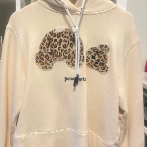 Beige hoodie från Palm Angels - Säljer en beige hoodie från Palm Angels med ett coolt leopardmönstrat nallemotiv på framsidan. Hoodien har en justerbar dragsko i huvan och är perfekt för en avslappnad stil.