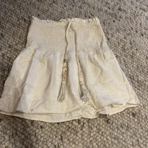 Beige kjol med knytband - Charmig beige kjol med elastisk midja och knytband. Kjolen har en smockad design upptill och dekorativa detaljer i tyget. 