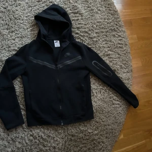 Svart hoodie från Nike - Snygg svart hoodie från Nike med dragkedja och huva. Perfekt för en casual look eller träning. Den har en diskret Nike-logga på bröstet och är tillverkad i ett mjukt material för extra komfort. Nyskick. Nypris 1199:- 