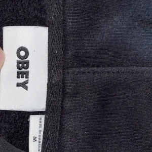 Svart hoodie från Obey - Säljer en svart hoodie från Obey med logga på bröstet. Den har en klassisk design med justerbar huva och en stor magficka. Perfekt för en avslappnad stil.
