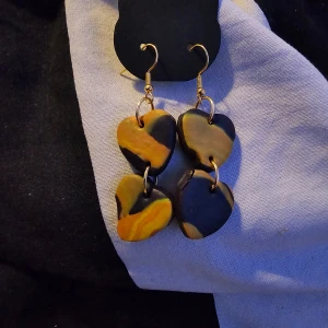 Flerfärgade hjärtformade örhängen - Unika örhängen med hjärtformade hängen i svart, gul och orange. Perfekta för att ge en färgglad touch till din outfit. De har en enkel krok för enkel användning. Guldpläterade.