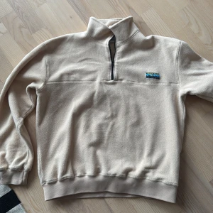 Beige fleece tröja från Junkyard - Säljer en beige fleece tröja från Junkyard med halv dragkedja och en Urban Tears patch på bröstet. Perfekt för kyliga dagar med sin mjuka och varma känsla. Tröjan har långa ärmar och ribbade muddar.