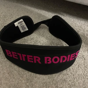 Svart träningsbälte från Better Bodies - Säljer ett svart träningsbälte från Better Bodies med rosa text. Perfekt för att ge extra stöd vid tunga lyft. Bältet är brett och justerbart för optimal passform.     aldrig använd