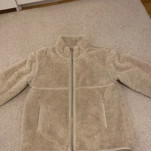 Beige teddyjacka från H&M - Mysig beige teddyjacka från H&M med dragkedja framtill. Perfekt för kyliga dagar med sin fluffiga och varma design. Jackan har långa ärmar och två praktiska fickor. Stor i storleken 