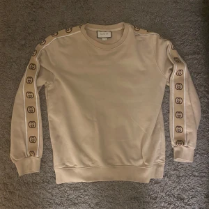 Beige sweatshirt från Gucci - Snygg beige sweatshirt från Gucci med deras ikoniska logotyp längs ärmarna. Tröjan har en klassisk rund hals och ribbade muddar vid ärmslut och nederkant. Perfekt för en stilren look.