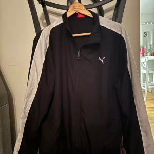 Svart träningsjacka från Puma - Säljer en svart träningsjacka från Puma med grå detaljer. Jackan har en dragkedja framtill och långärmade ärmar. Perfekt för sportiga aktiviteter eller som en lätt överdragströja. Liten fläck som går att ta bort vid tvätt. 