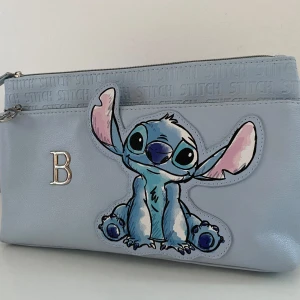 Blå sminkväska med Stitch Disney  - Säljer en söt blå necessär med ett stort Stitch-motiv på framsidan. Väskan har en dragkedja upptill och ett dekorativt B-emblem. Perfekt för alla som älskar Disney och vill ha en unik accessoar.